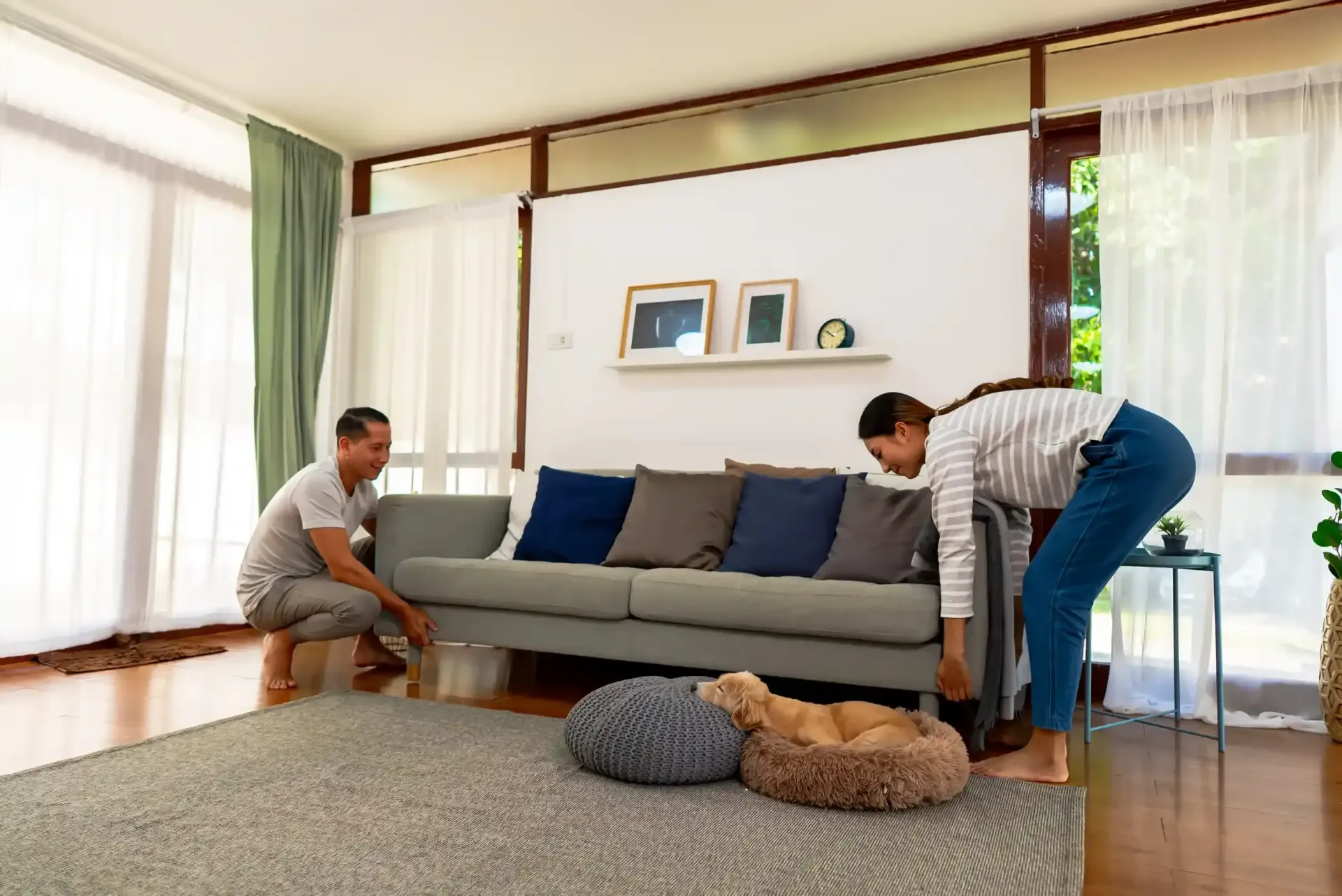 En un luminoso salón, una pareja levanta un sofá gris para limpiar debajo