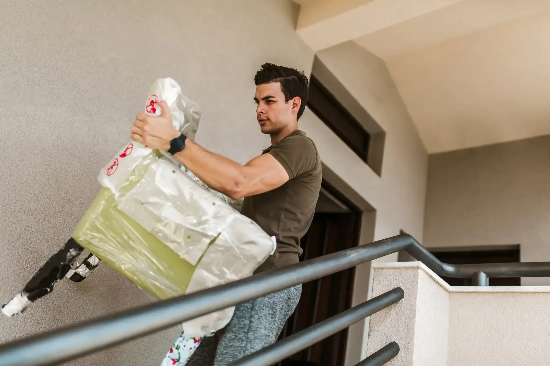 Una persona que lleva una camiseta sube un mueble embalado por una escalera mientras se sujeta a la barandilla.
