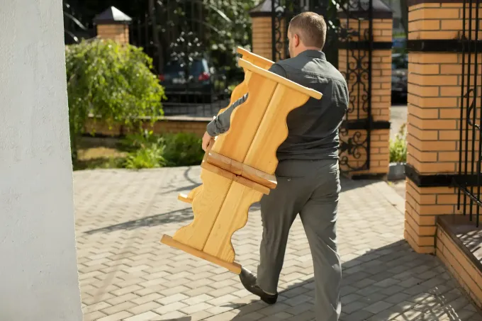 Una persona camina al aire libre por un sendero pavimentado mientras lleva un banco de madera sobre su hombro.