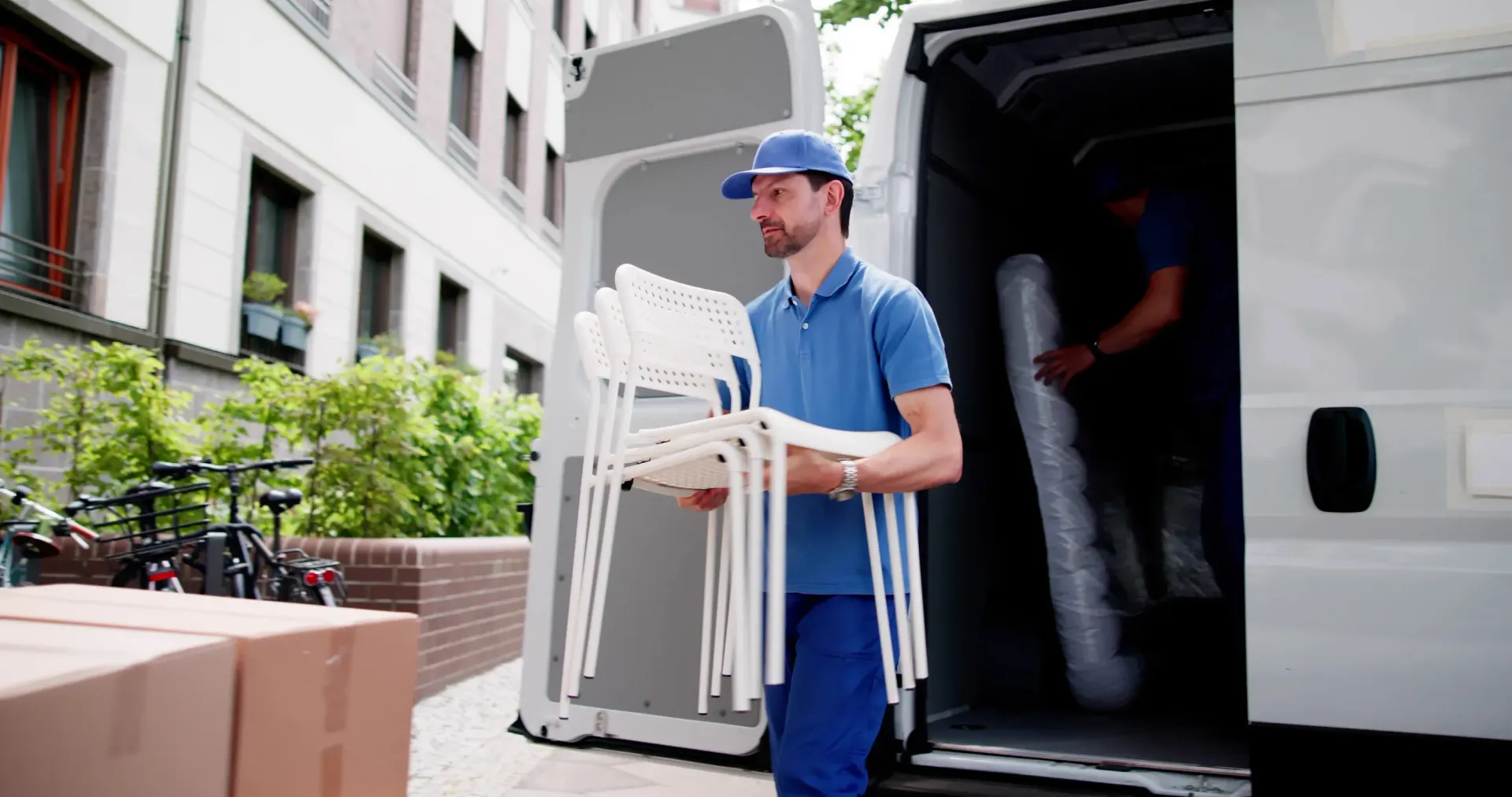 Un repartidor con uniforme azul lleva sillas blancas apiladas hacia una furgoneta aparcada en una calle de la ciudad.