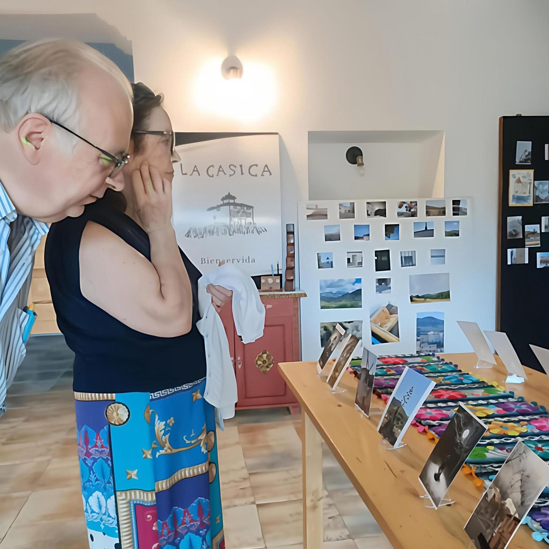 Dos personas mirando postales y fotos expuestas en una mesa en una galería o tienda luminosa.