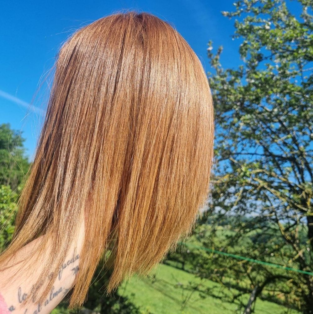 Brazo de mujer sosteniendo una peluca de color cobre, contra un cielo azul brillante y vegetación.