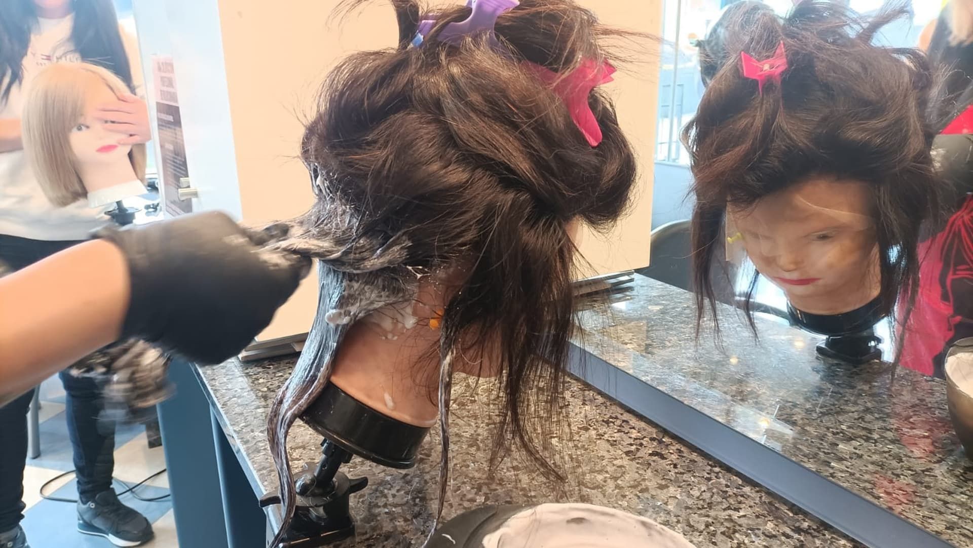 Una persona con guantes negros aplica tinte para el cabello al cabello oscuro de un maniquí en un salón.