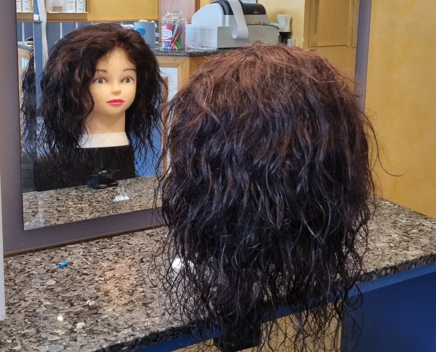 Cabeza de maniquí con cabello oscuro y mojado rizado frente a un espejo, reflejando el rostro del maniquí.