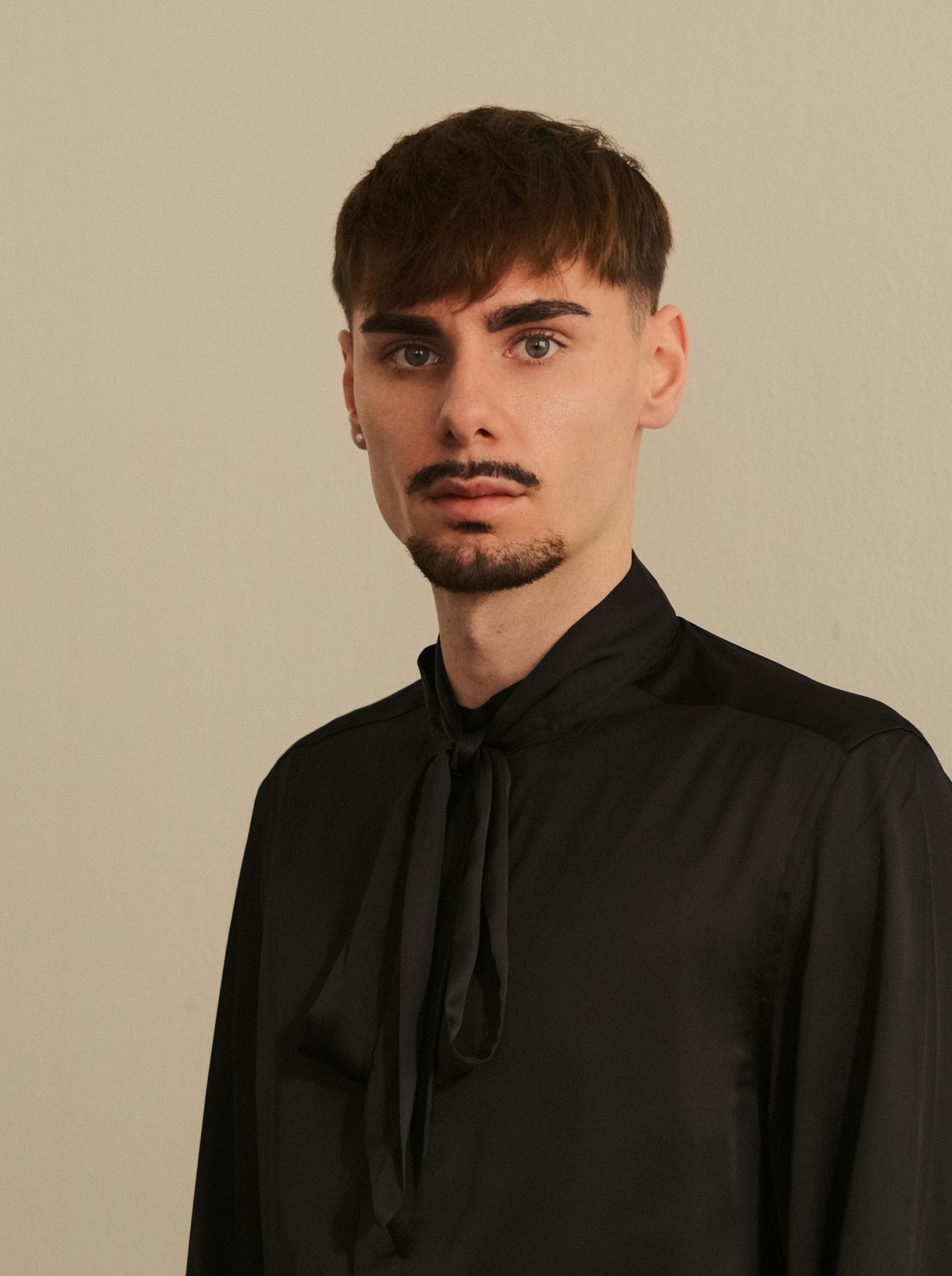 Un joven con bigote, vestido con una camisa negra y corbata, mira a la cámara.