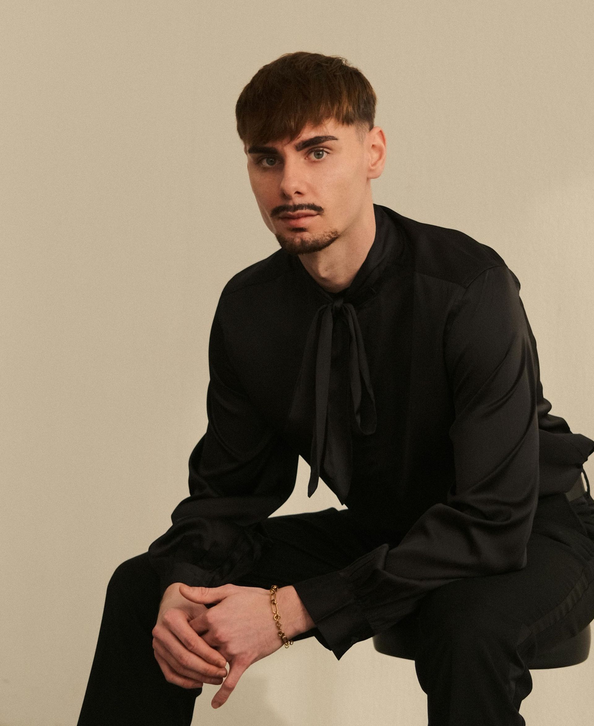 Hombre vestido de negro, sentado. Cabello castaño, bigote, camisa con lazo, brazalete dorado y fondo beige.