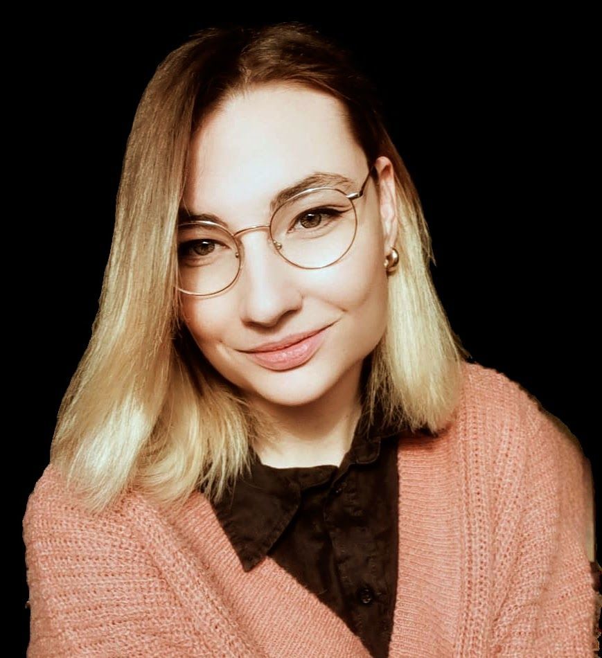 Eine Person mit blonden, schulterlangen Haaren und runder Brille lächelt vor schwarzem Hintergrund und trägt eine rosa Strickjacke.