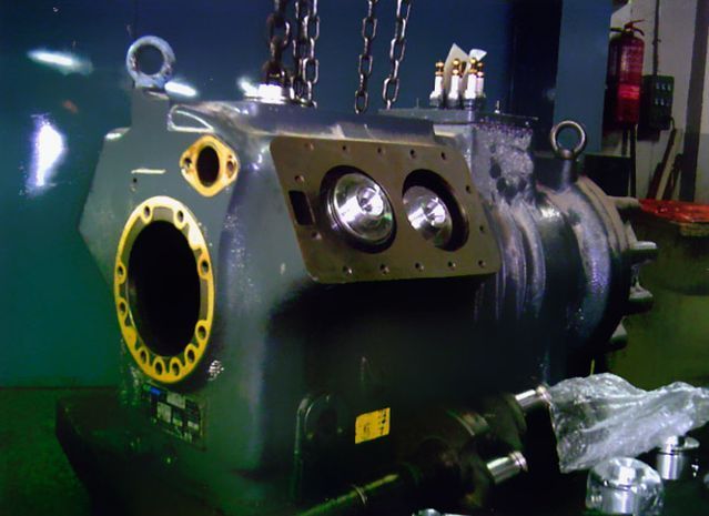 Caja de engranajes industrial grande, de metal gris, con accesos abiertos. Colgada de cadenas.