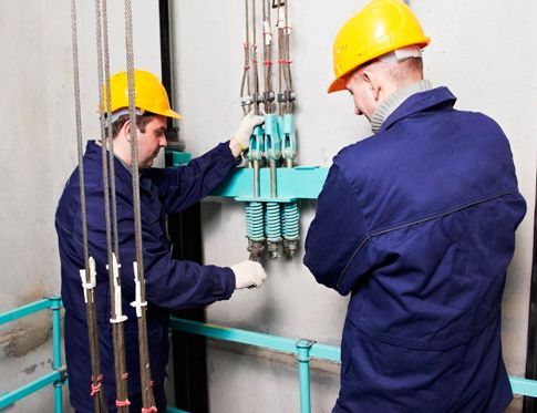 Dos trabajadores con overoles azules y cascos inspeccionan los cables y la maquinaria del ascensor.