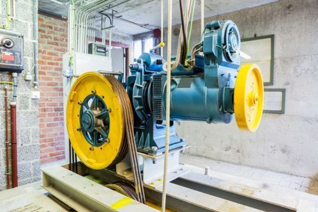 Motor de ascensor y sistema de poleas. Motor azul con ruedas amarillas, cables colgando. Paredes de hormigón y ladrillo.