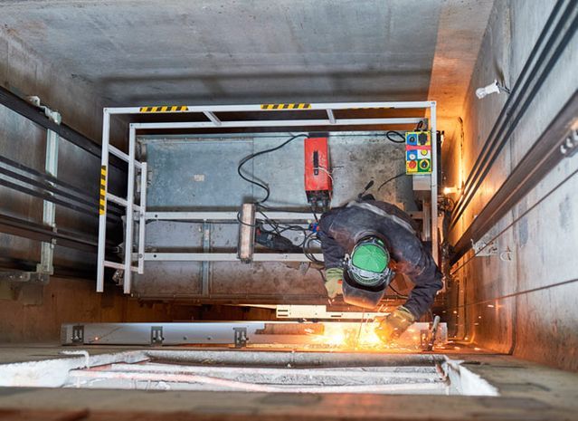 Un trabajador suelda metal en el hueco de un ascensor. Saltan chispas; hay una jaula protectora encima. Lleva casco de seguridad.