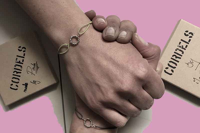Hände halten goldene Armbänder mit zwei „Cordels“-Boxen auf rosa Hintergrund.
