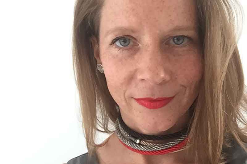 Frau mit heller Haut, rotem Lippenstift und einem geflochtenen Halsband lächelt in die Kamera.