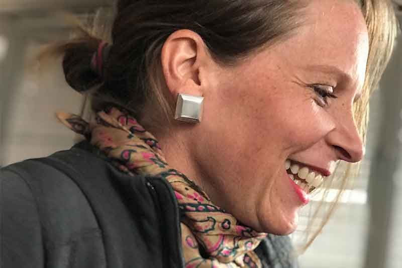 Frau mit heller Haut, lächelnd, mit quadratischen Ohrringen und einem gemusterten Schal.