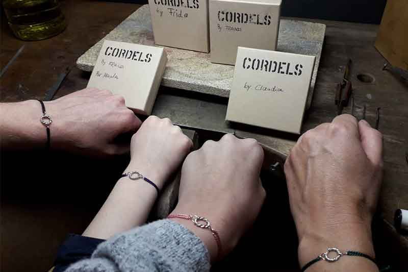 Vier Personen tragen passende Armbänder und beige Schachteln mit der Aufschrift „CORDELS“ in einer Werkstattumgebung.