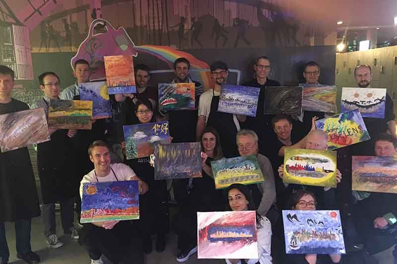 Eine Gruppe von Menschen hält selbst gemalte bunte Bilder hoch. Drinnen, vor einem Wandgemälde und einem Regenbogen.