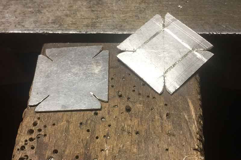 Zwei quadratische Metallstücke mit abgeschnittenen Ecken, eines flach, das andere zu einer 3D-Form gefaltet, auf einer hölzernen Arbeitsfläche.