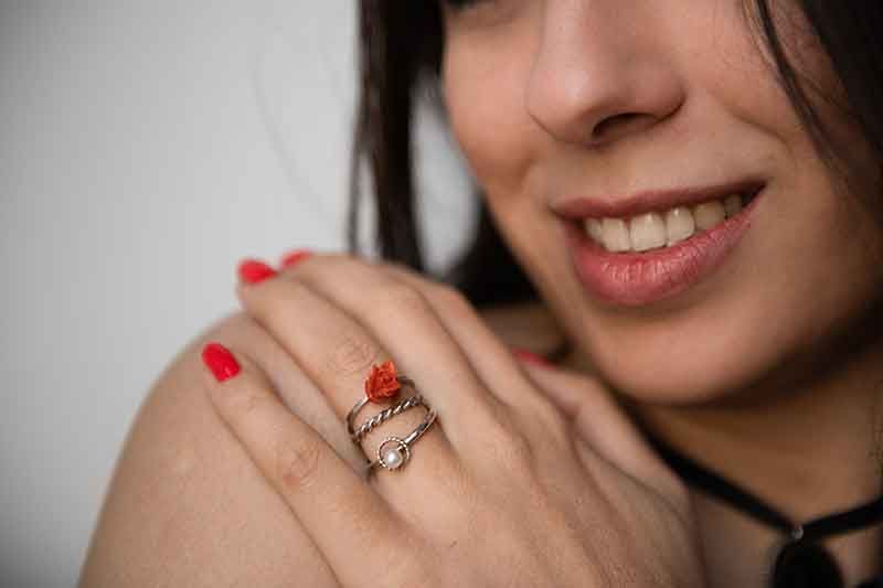 Frau trägt einen Ring mit roter Koralle, lächelt und berührt ihre Schulter; rote Nägel, schwarzes Halsband.
