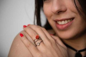 Frau trägt einen Ring mit roter Koralle, lächelt und berührt ihre Schulter; rote Nägel, schwarzes Halsband.