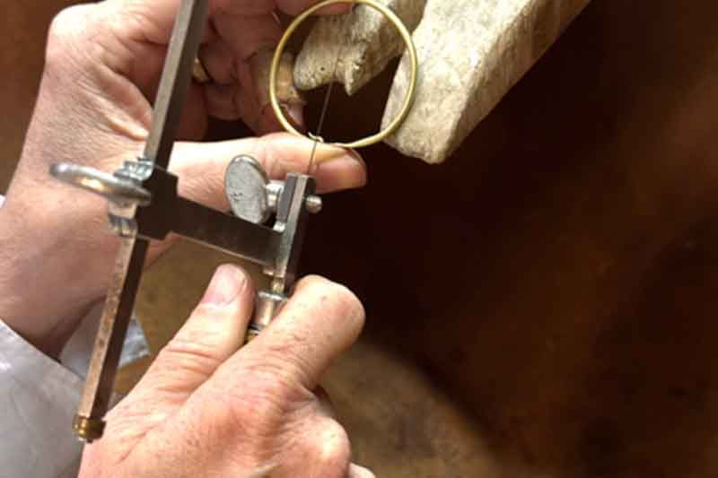 Hände, die mit einer Juweliersäge einen Goldring schneiden, im Hintergrund eine hölzerne Werkbank.