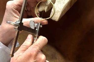 Hände, die mit einer Juweliersäge einen Goldring schneiden, im Hintergrund eine hölzerne Werkbank.