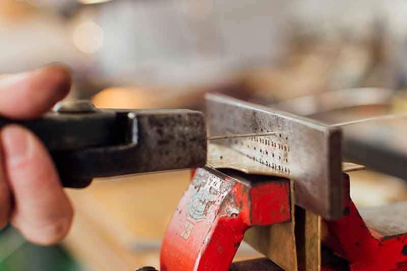 Eine Person stanzt mit einem Handwerkzeug Löcher in eine Metallplatte, die in einem roten Schraubstock eingespannt ist.