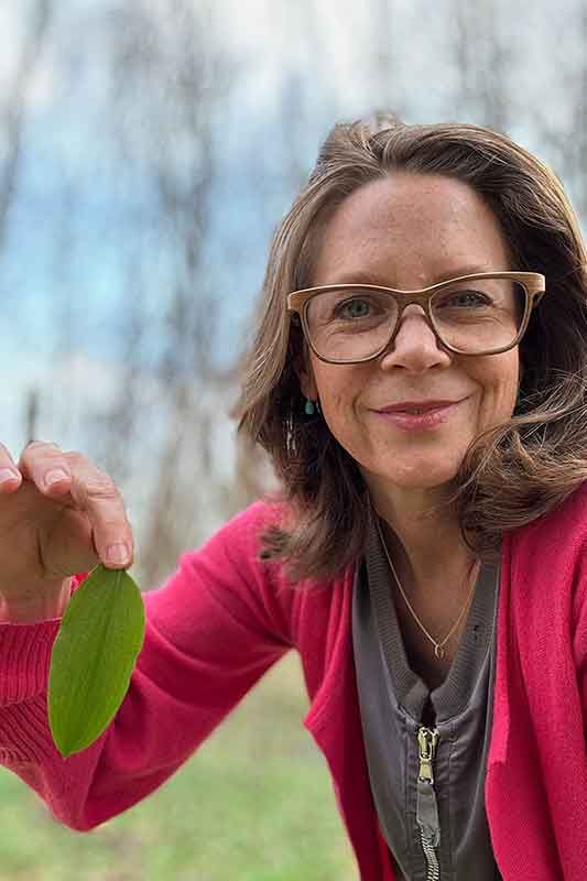 Frau mit Brille, die ein grünes Blatt hält, lächelt im Freien, trägt einen rosa Pullover und ein graues Oberteil.
