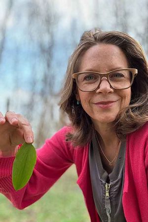 Frau mit Brille, die ein grünes Blatt hält, lächelt im Freien, trägt einen rosa Pullover und ein graues Oberteil.