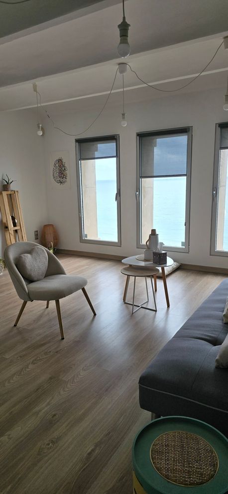 Un escritorio de madera con una taza de café y un ordenador portátil, con vistas a un soleado paisaje marino a través de dos grandes ventanales.