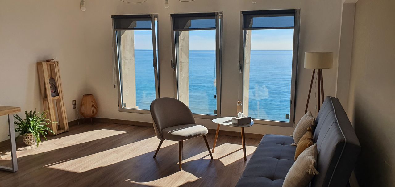 Un escritorio de madera con una taza de café y un ordenador portátil, con vistas a un soleado paisaje marino a través de dos grandes ventanales.
