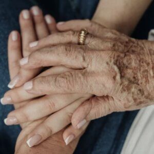 Primer plano de una persona con cambios en la piel relacionados con la edad, que sostiene la mano de otra persona con una tez tersa y bien cuidada.