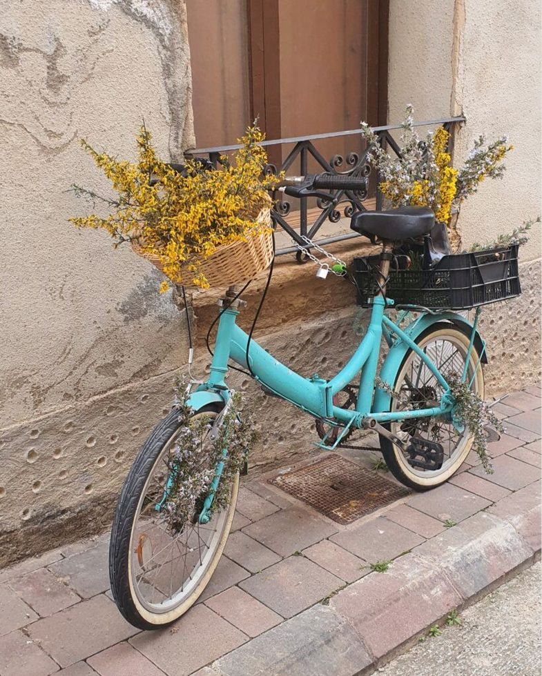 Una bicicleta turquesa decorada con flores amarillas en la cesta delantera y en una caja trasera, aparcada en una acera de piedra.