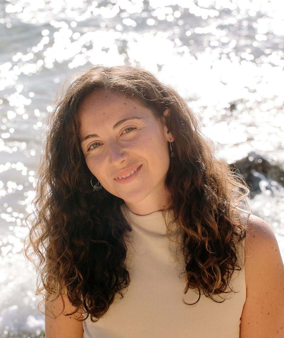 Persona sonriente con cabello rizado y blusa beige, de pie cerca de agua cristalina bajo la luz del sol.
