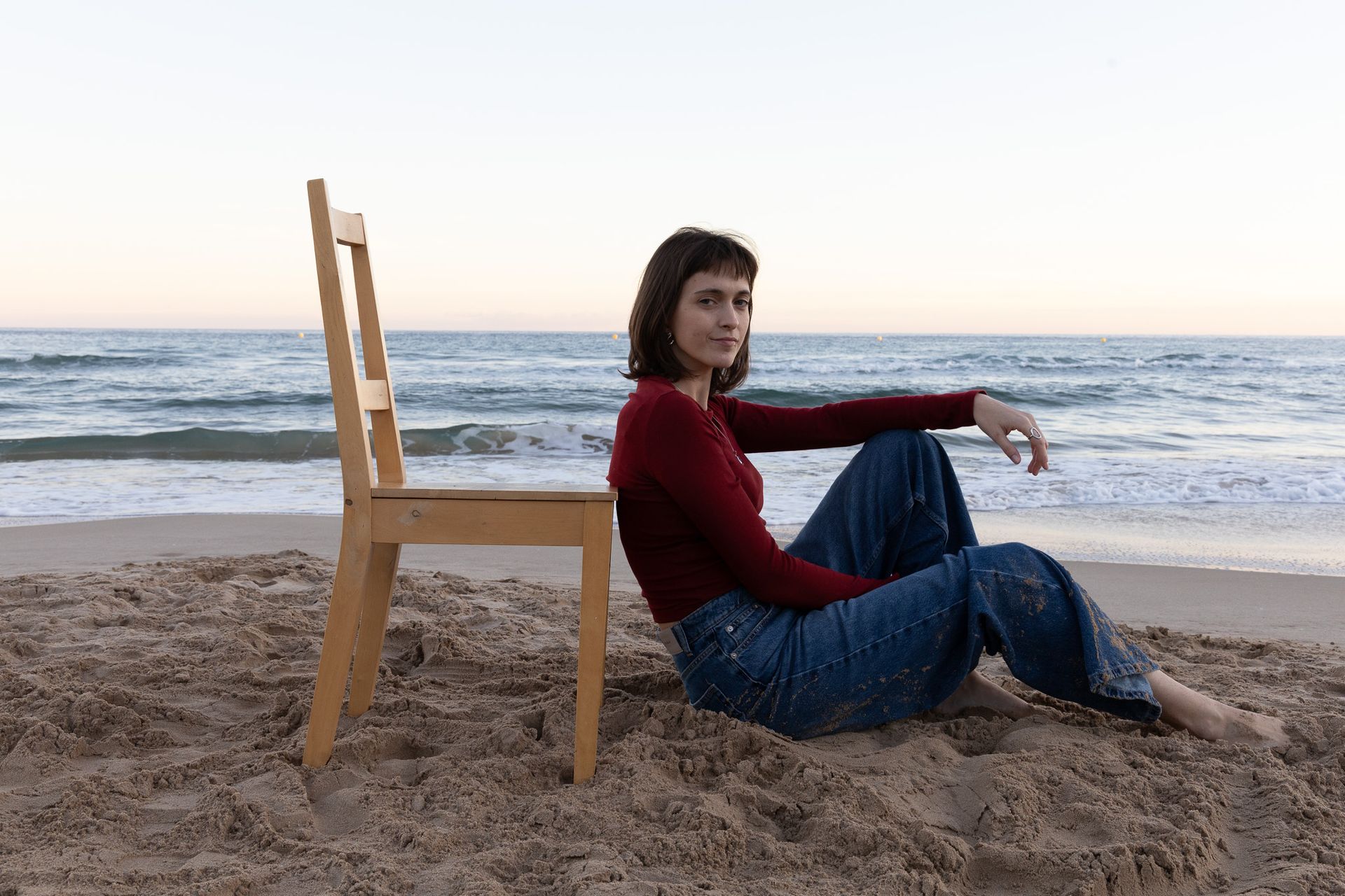 Una persona sentada en una playa de arena junto a una silla de madera, mirando al océano al atardecer.