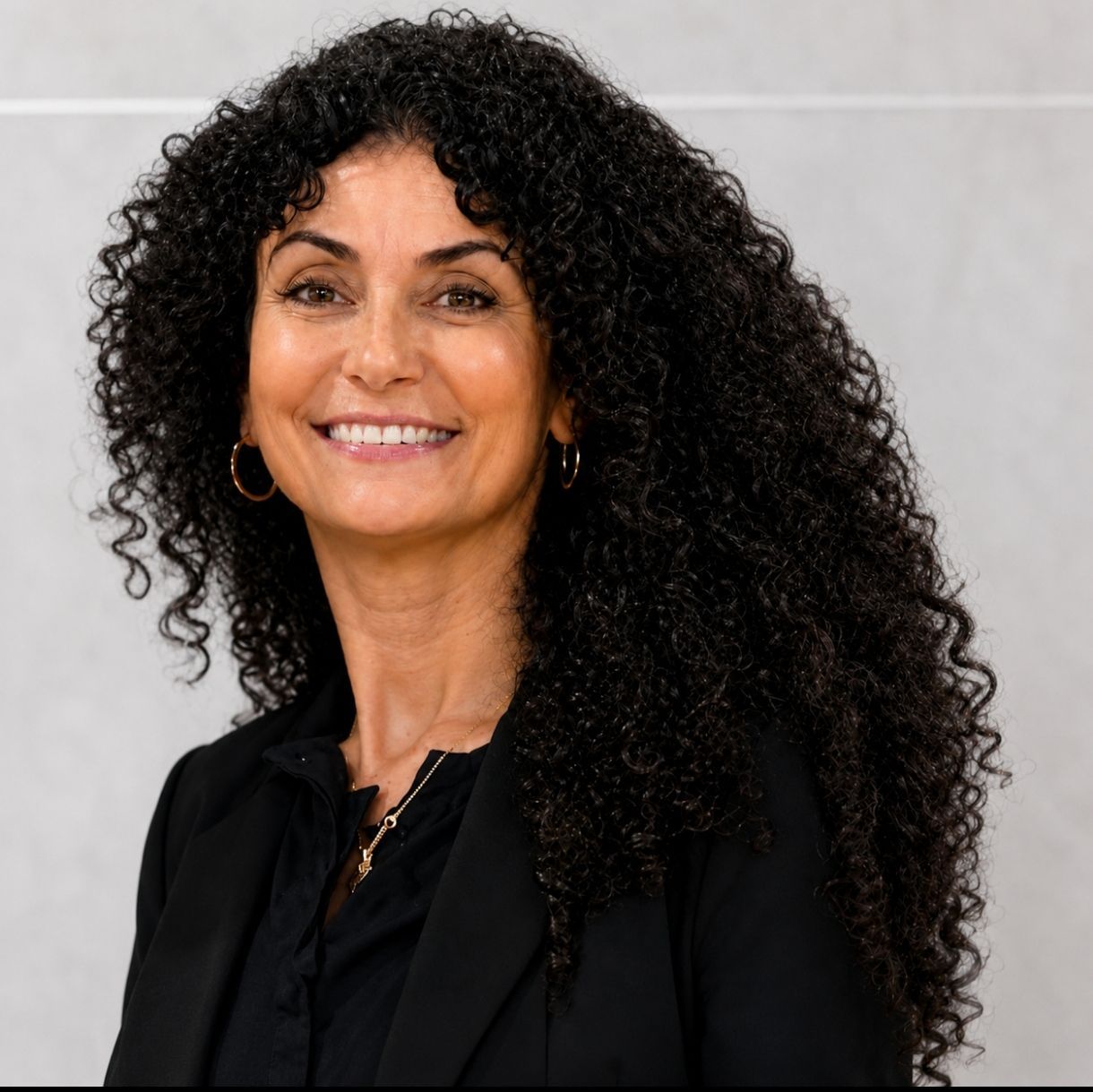 Mujer sonriente con cabello largo y rizado negro, vestida de negro sobre un fondo gris claro.