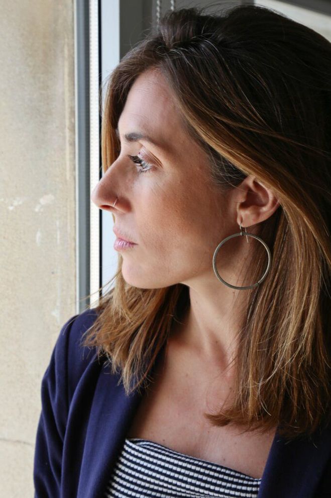 Una persona con una chaqueta azul y una camiseta de rayas mira por la ventana con expresión neutra, luciendo grandes pendientes de aro.