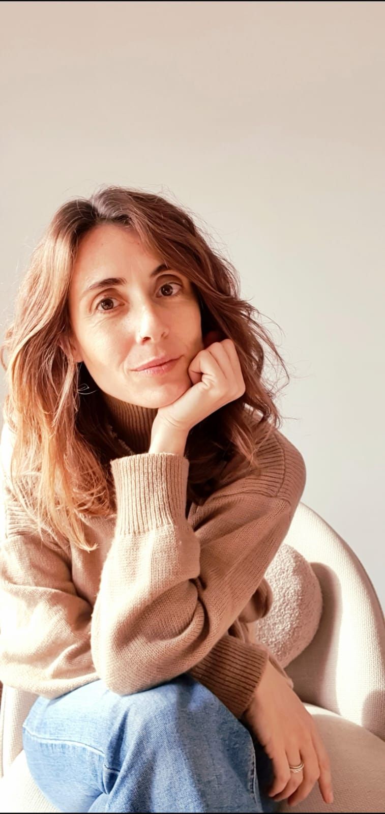 Una persona con cabello castaño ondulado apoya la barbilla en la mano, vestida con un suéter color canela y jeans azules, recostada contra una pared clara.