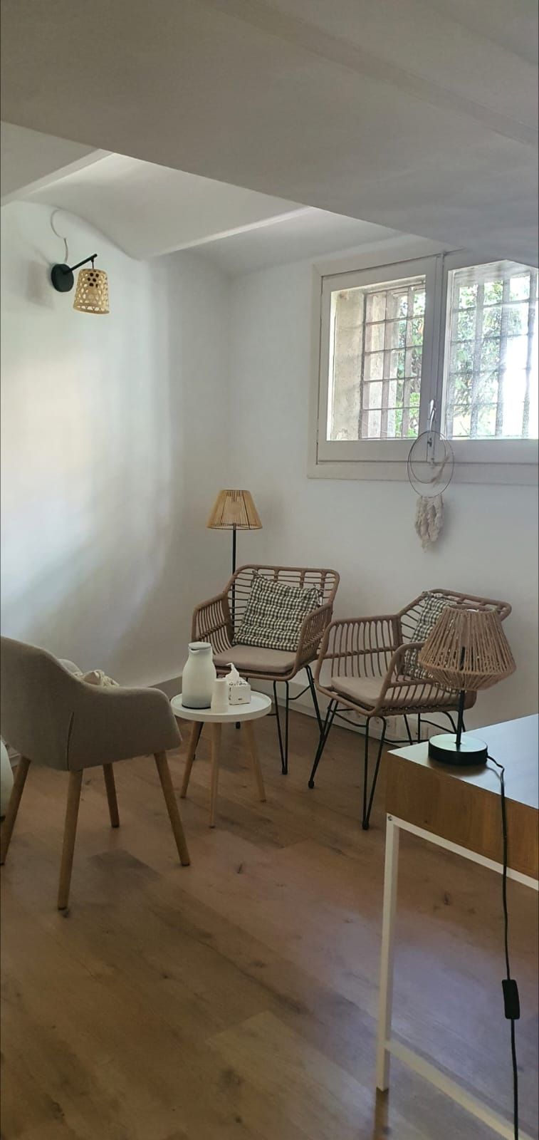 Una habitación acogedora con suelos de madera clara, tres sillas de mimbre, una mesita auxiliar y una ventana con reja metálica.