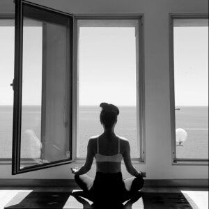 Una persona se sienta en postura meditativa sobre una esterilla de yoga frente a unos grandes ventanales con vistas al mar, capturada en silueta.