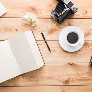 Un cuaderno abierto, un bolígrafo, un papel arrugado, una taza de café y una cámara antigua sobre una superficie de madera clara.