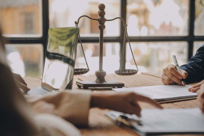 Una balanza de la justicia reposa sobre una mesa de madera entre dos personas que revisan documentos en un entorno de oficina profesional.