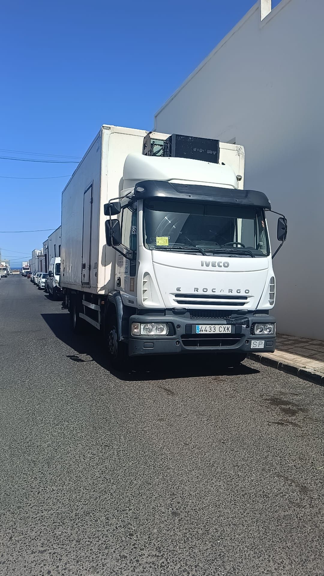 Camión refrigerado blanco estacionado en un camino de grava oscuro al lado de un edificio blanco bajo un cielo azul.