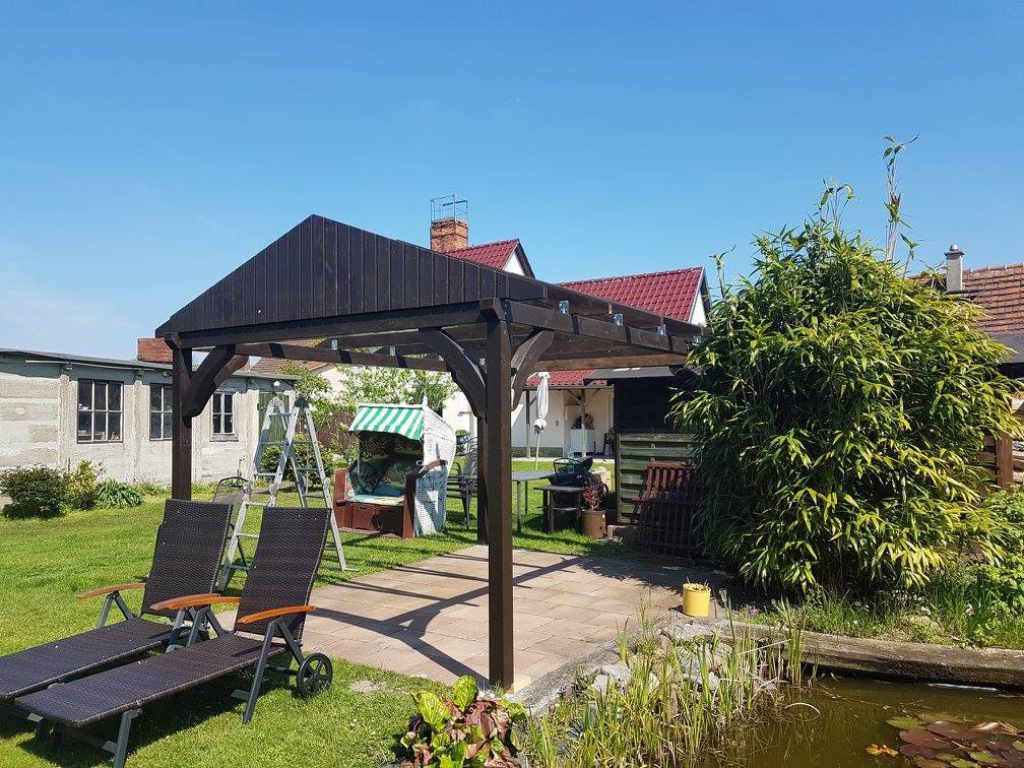 Foto einer Gartenüberdachung aus sehr dunklem Holz mit zwei Liegestühlen auf der Wiese davor