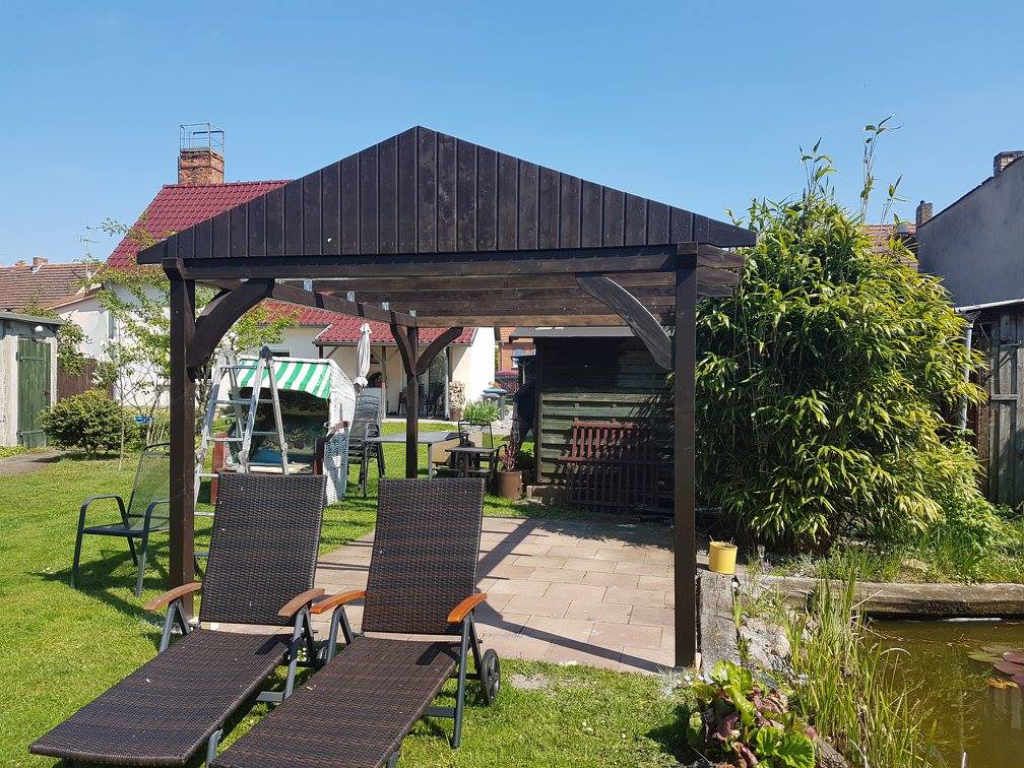 Frontbild einer Gartenüberdachung aus sehr dunklem Holz mit zwei Liegestühlen auf der Wiese davor