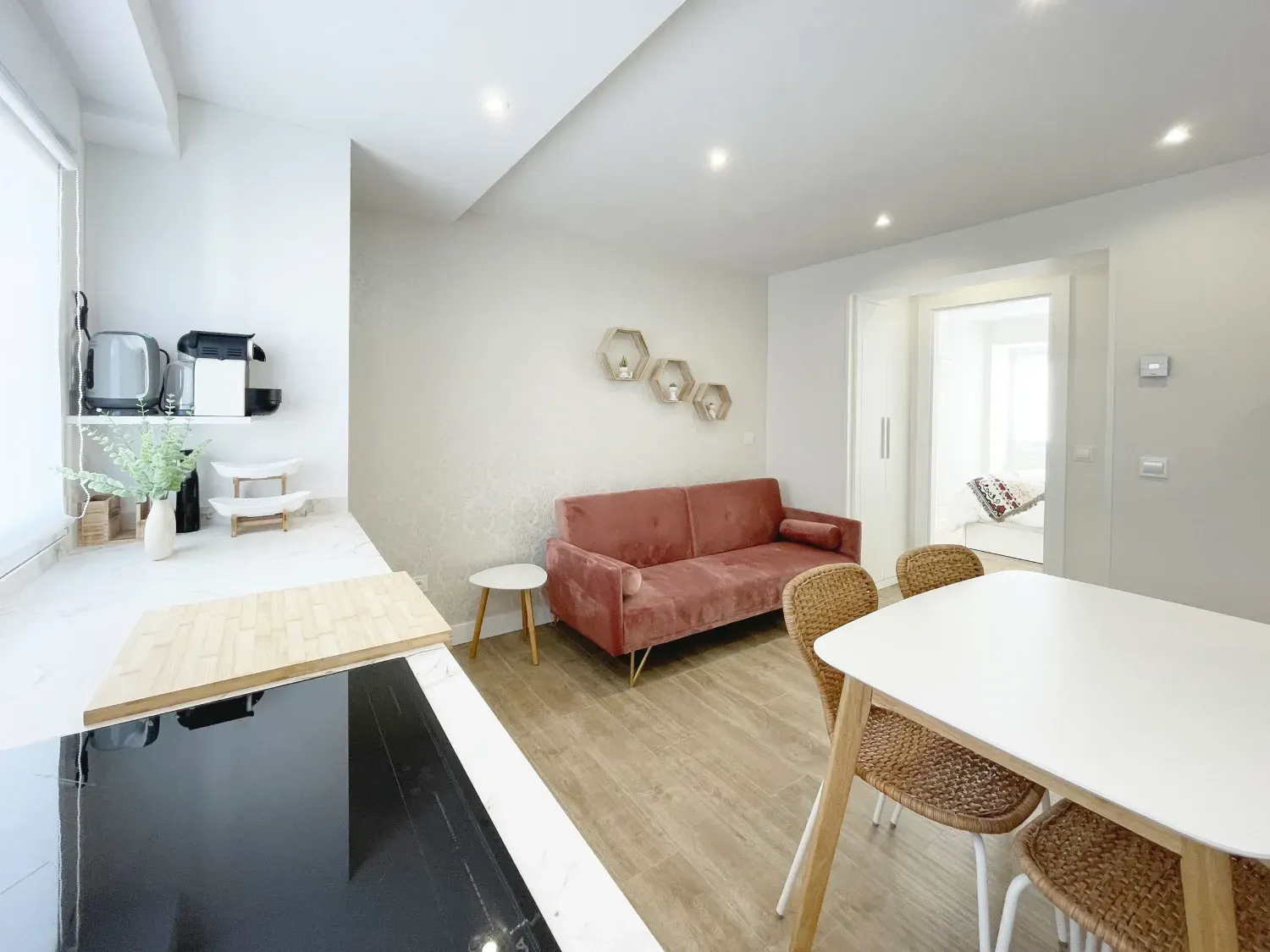 Una sala de estar pequeña y luminosa con encimera de cocina, mesa de comedor y un sofá rosa. Los suelos de madera