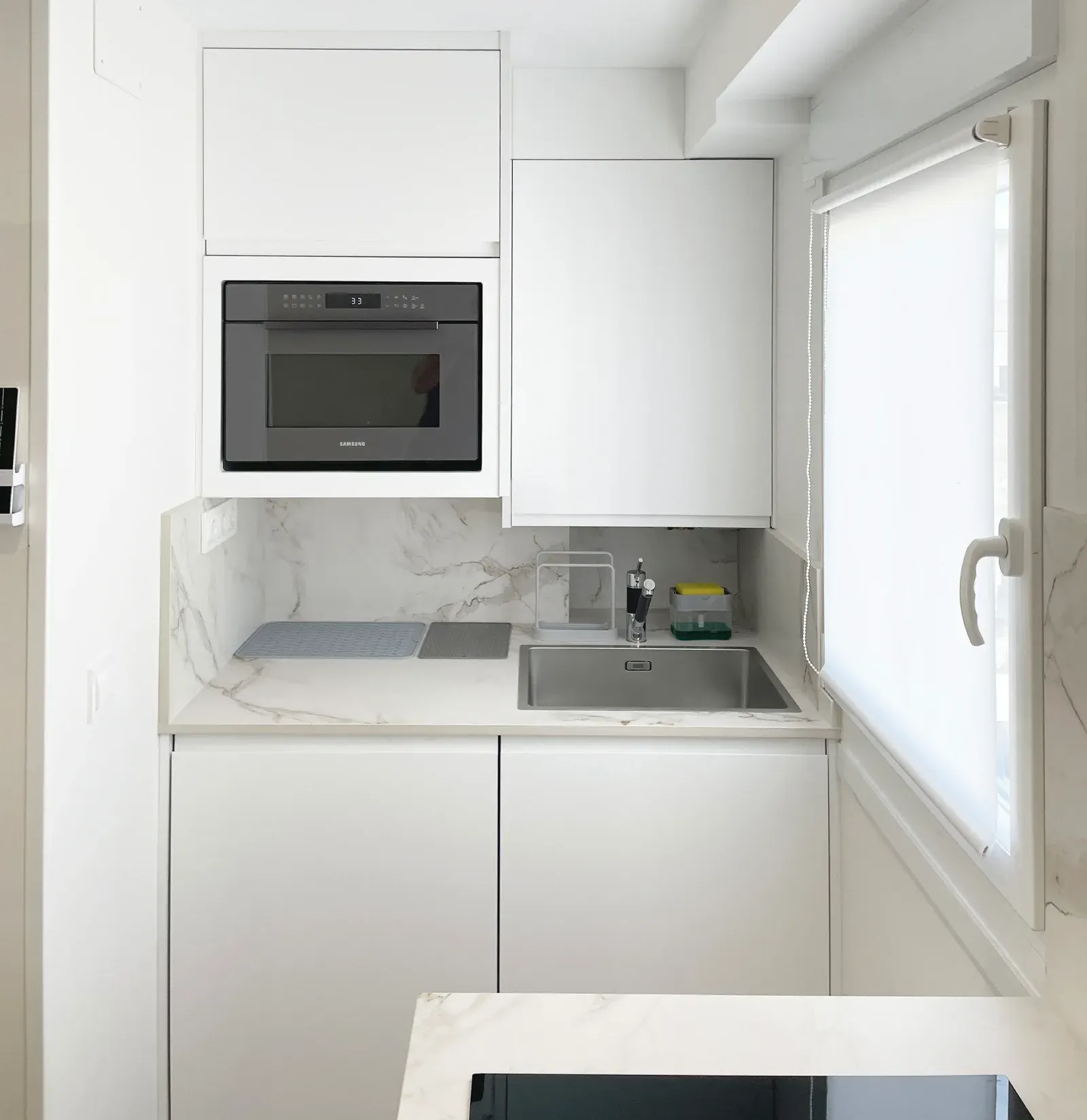 Cocina pequeña y blanca con microondas empotrado, fregadero y armarios. A la derecha hay una ventana con persiana blanca.