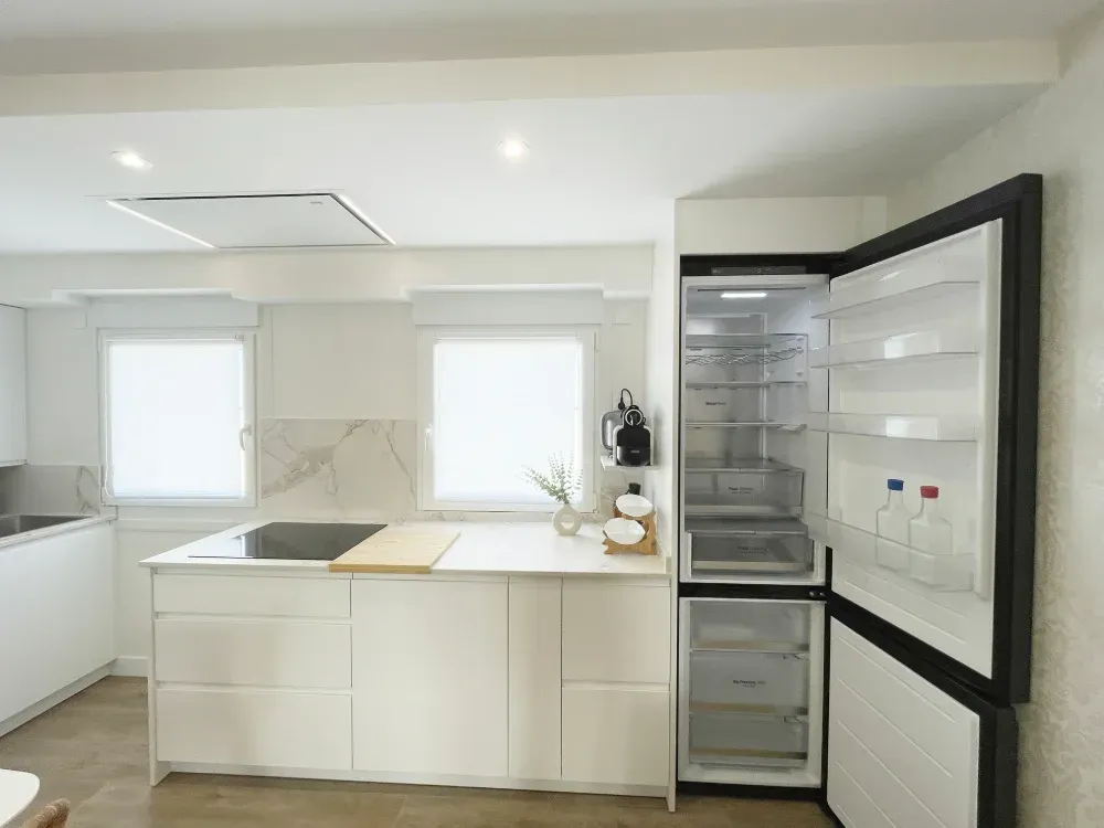 Cocina moderna con refrigerador abierto. Muebles y encimeras blancos. Dos ventanas con persianas.
