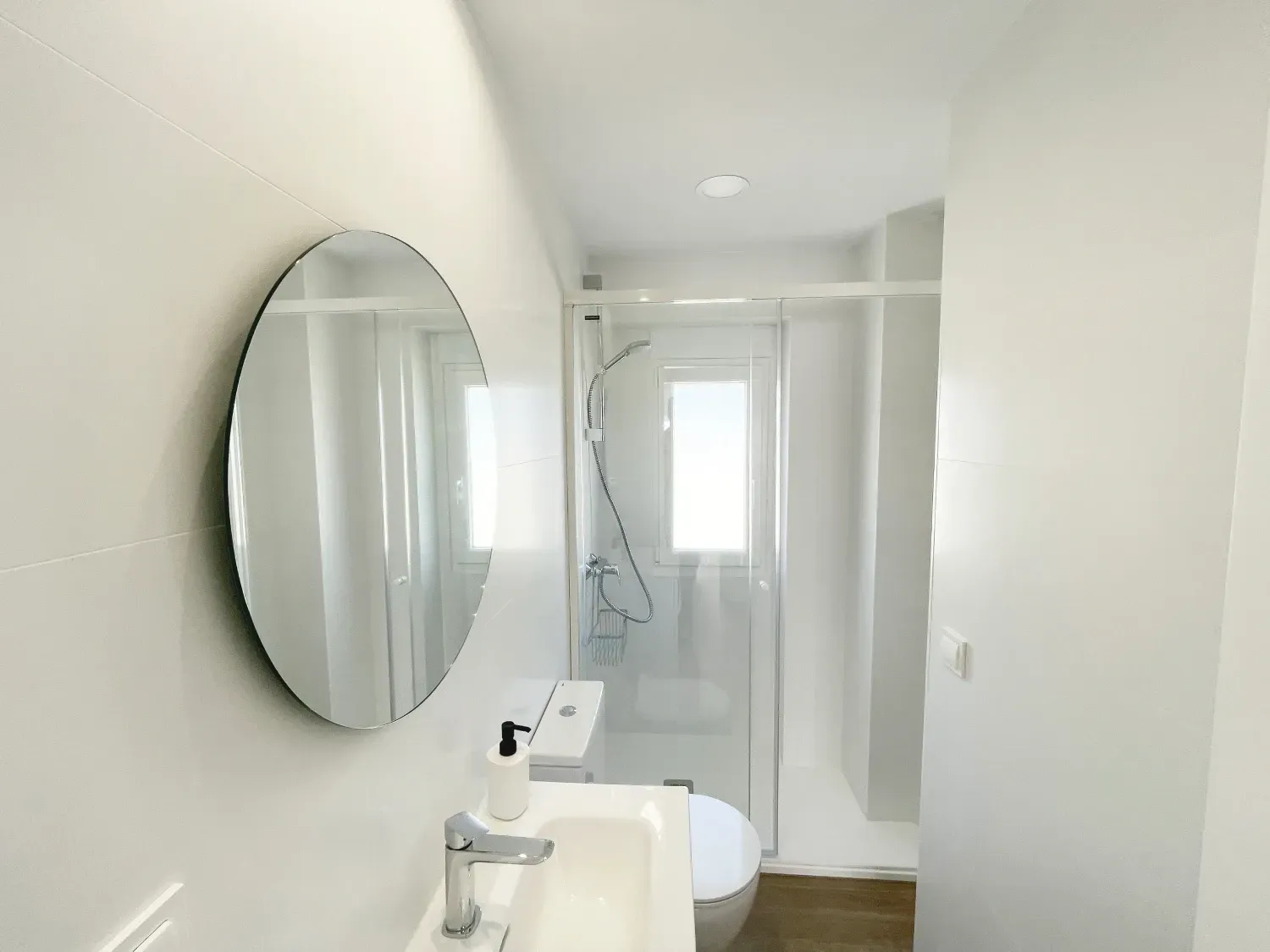 Pequeño baño blanco brillante con espejo redondo, lavabo, inodoro y ducha con mampara de cristal.