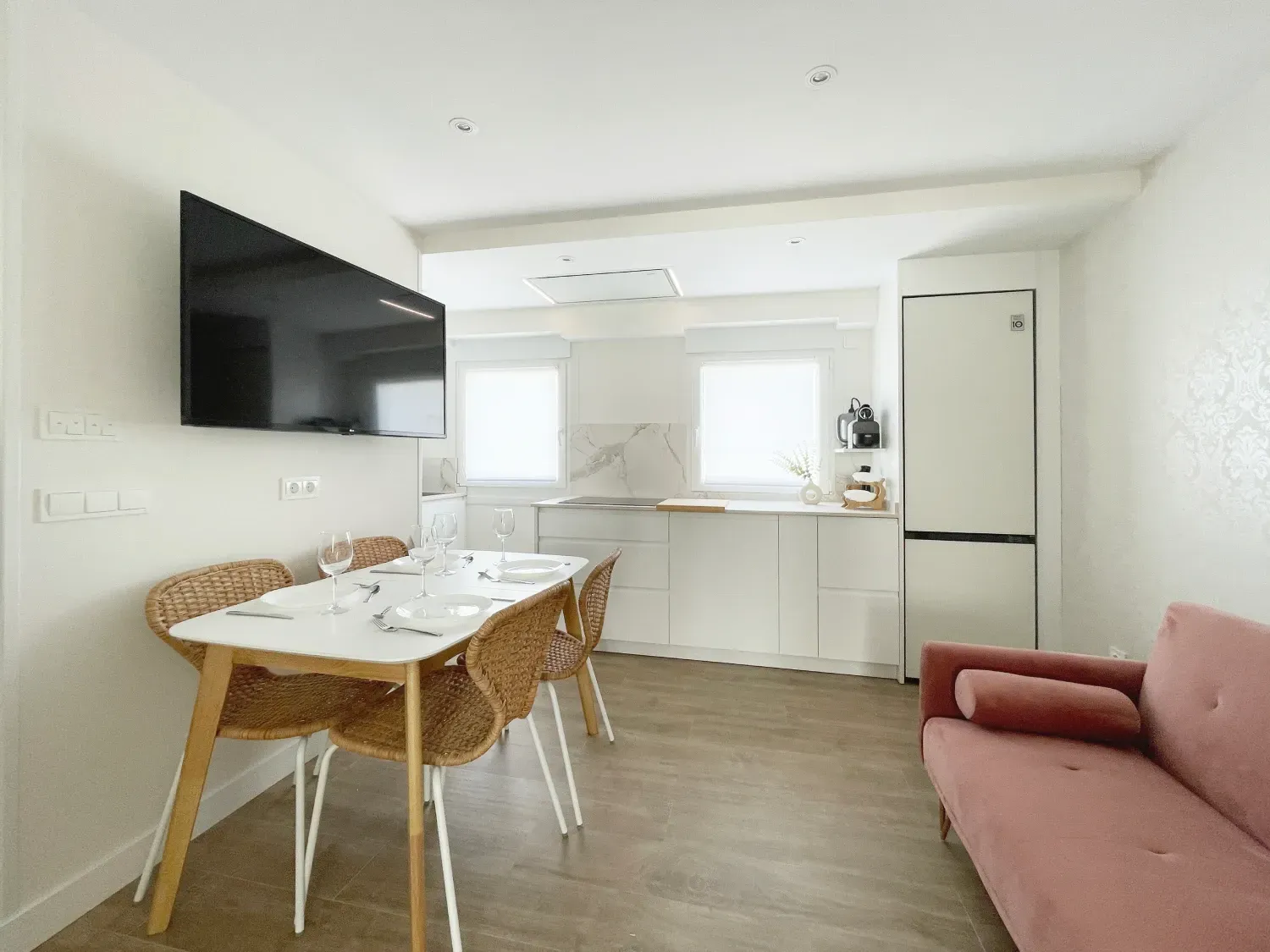 Cocina y comedor de concepto abierto. Paredes blancas, mesa de madera clara con sillas de mimbre y un sofá rosa.