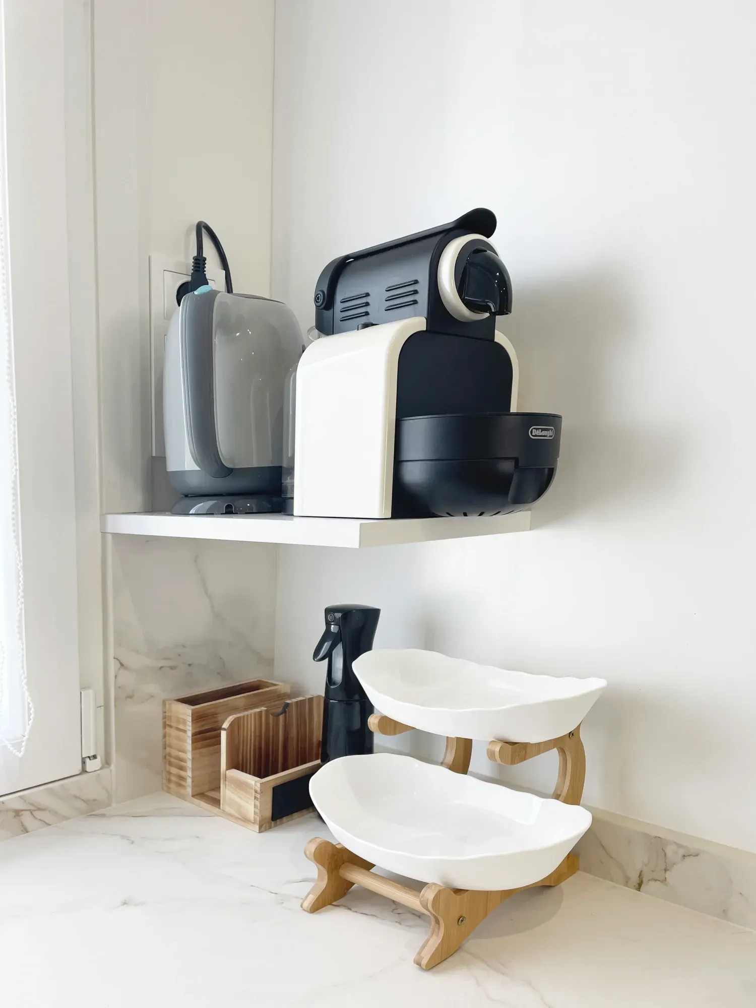 Un rincón de cocina con una cafetera y un hervidor de agua sobre un estante blanco. Debajo, dos tazones blancos